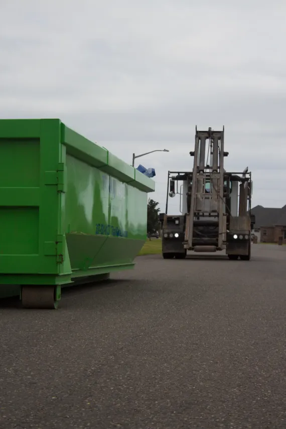 A green dumpster is being towed by a forklift