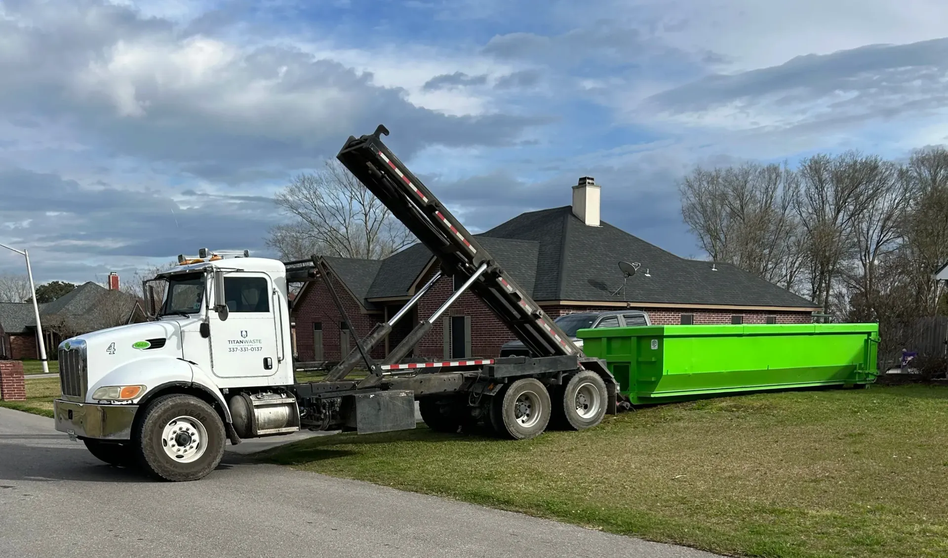 A dump truck is pulling a green dumpster in front of a house.