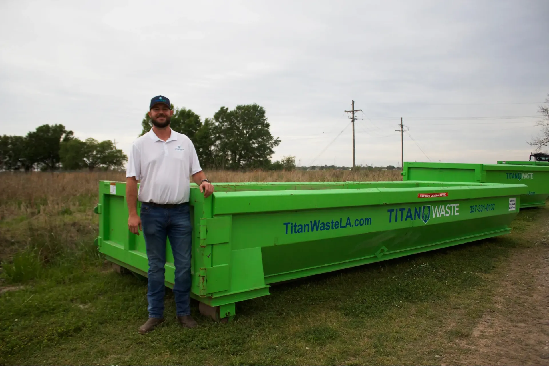 A man is standing next to a green dumpster in a field.