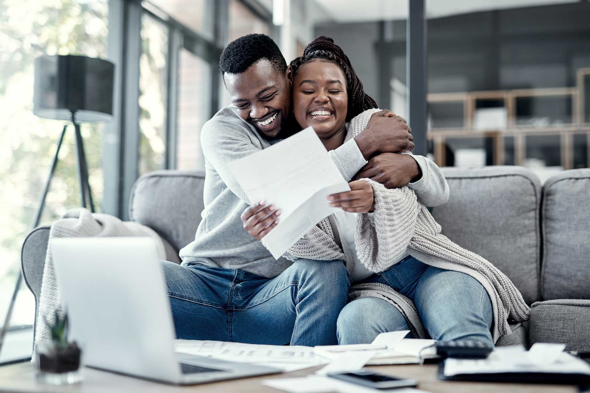 Couple on couch, smiling, hugging, reviewing paperwork with laptop and calculator in front of them.