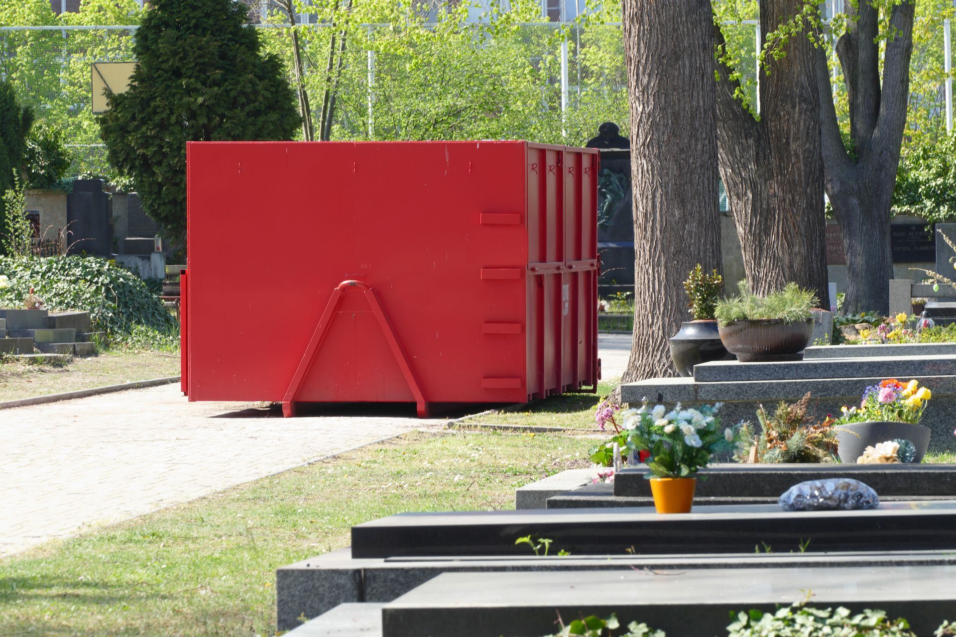 Bright red dumpster on a tree-lined park path beside flower beds