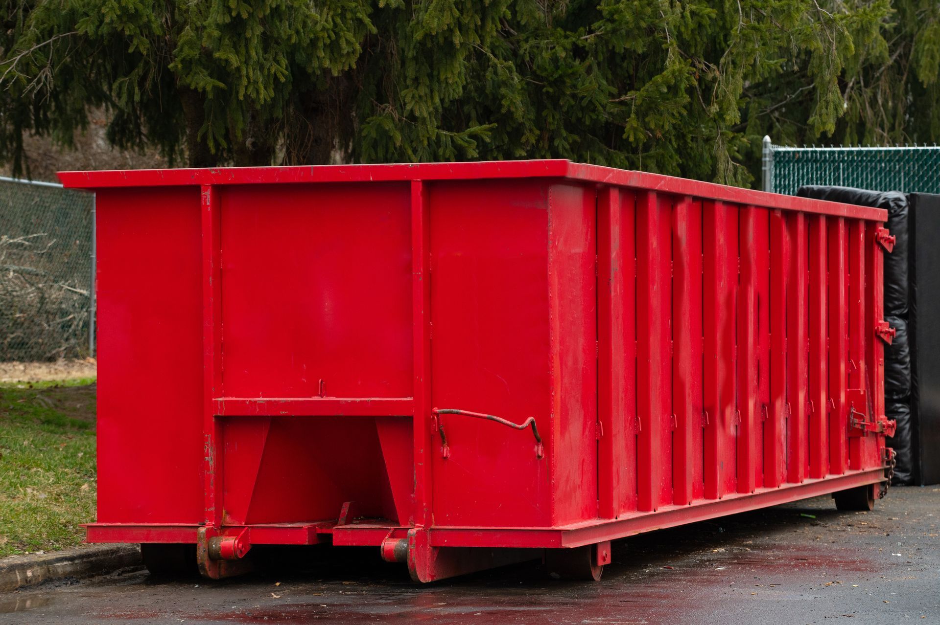 Red industrial dumpster container on a wet pavement outdoors, with trees in the background