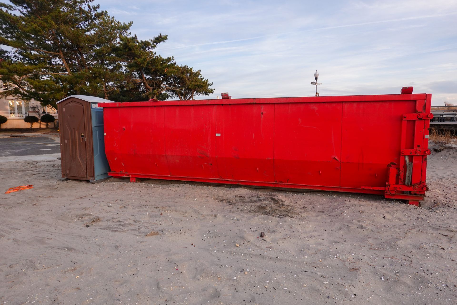 Overturned red dumpster on gravel lot near trees and parked cars