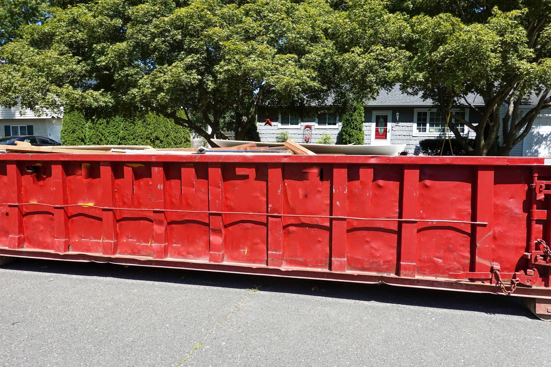Red metal dumpster on a street with trees and buildings in the background
