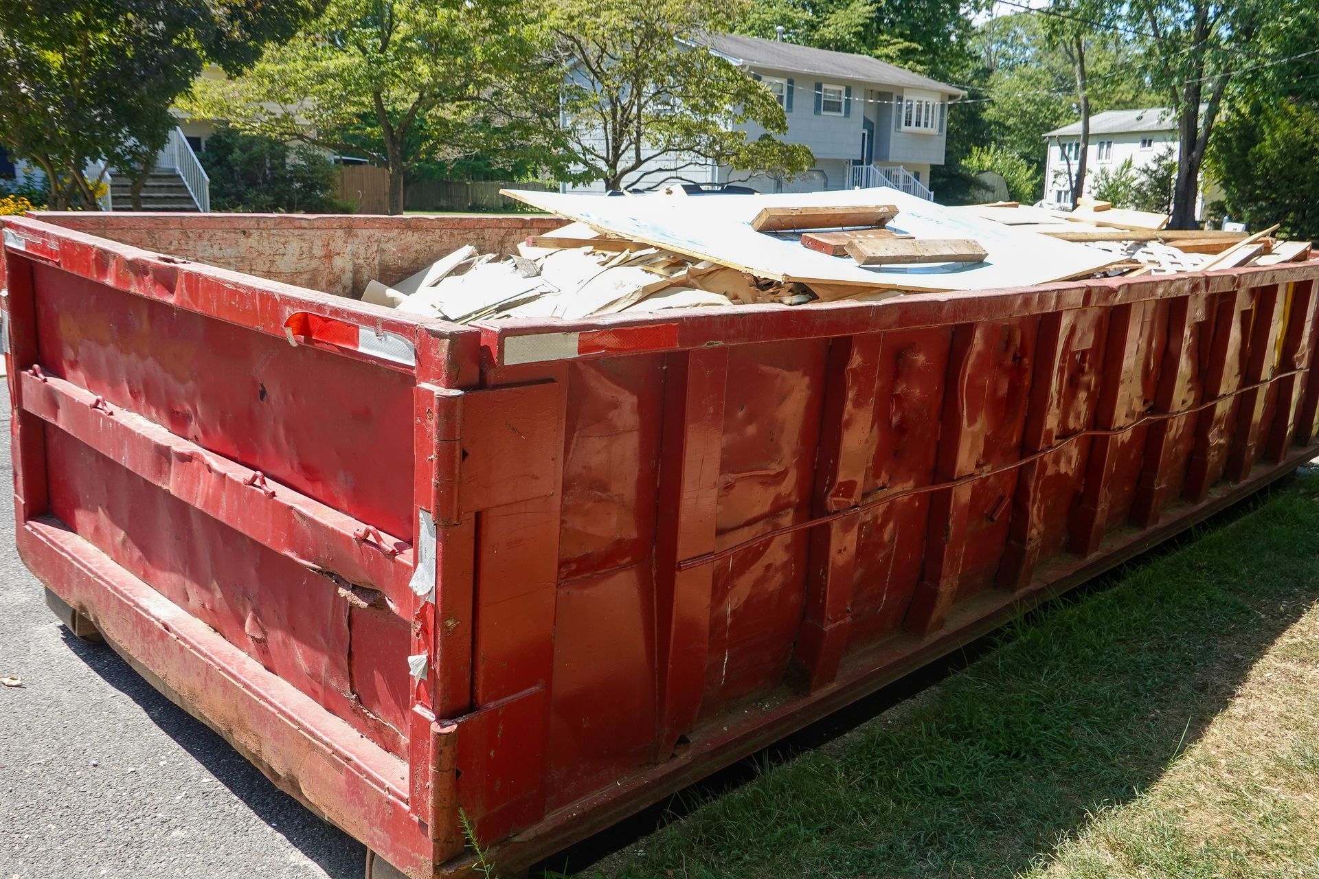 Large red metal dumpster filled with construction debris, sitting on grass near houses.