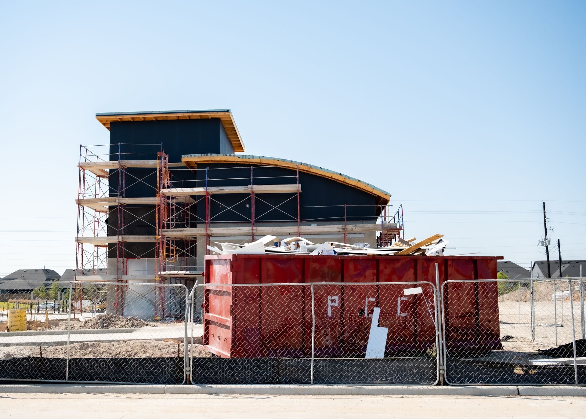 Partially built modern building beside a red dumpster behind a chain-link fence on a sunny day