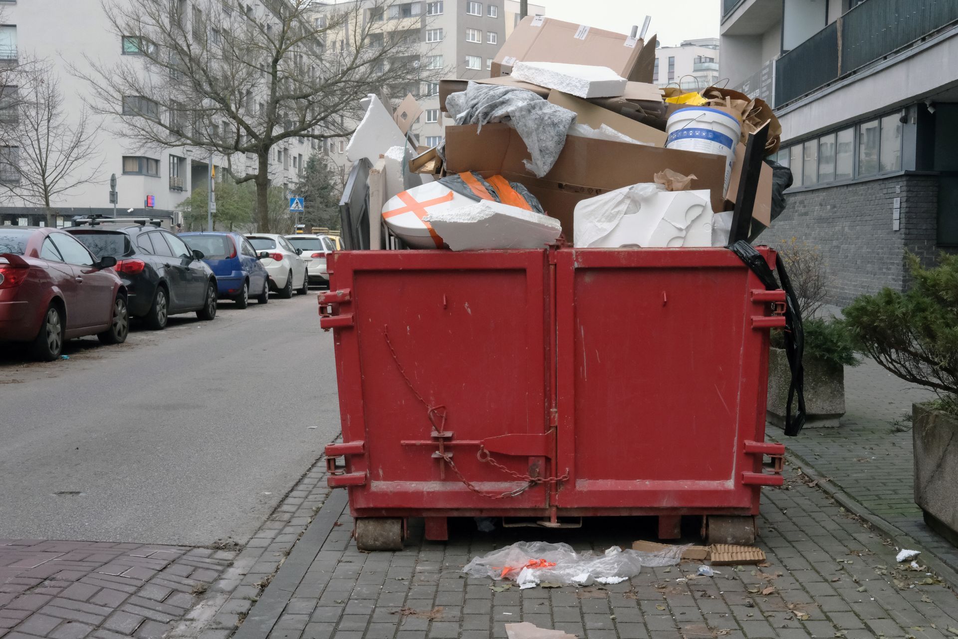 Overflowing red dumpster on a city street with trash piled on top and scattered on the pavement.