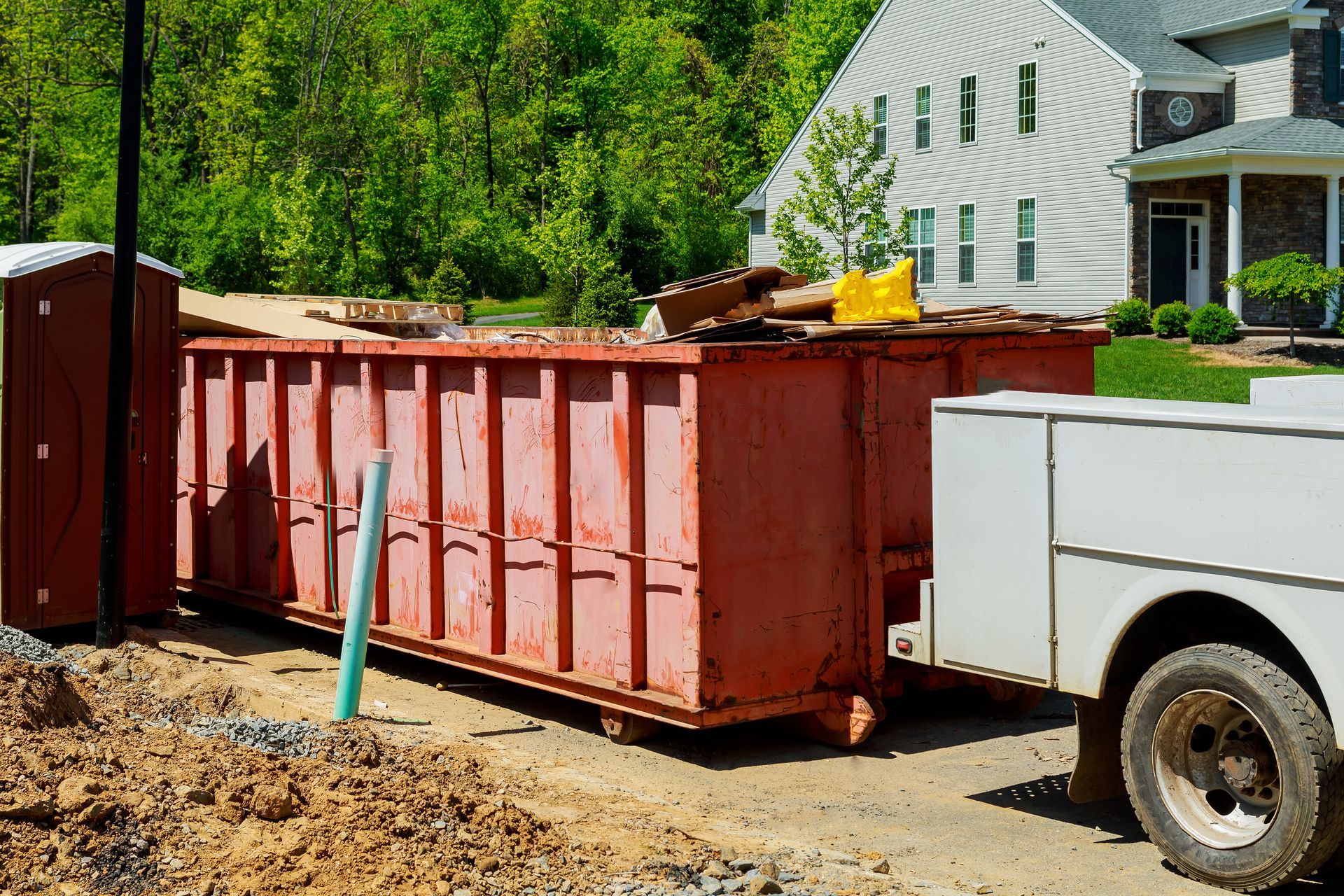 Red construction dumpster beside a white utility trailer in a muddy yard, with a house and trees in the background