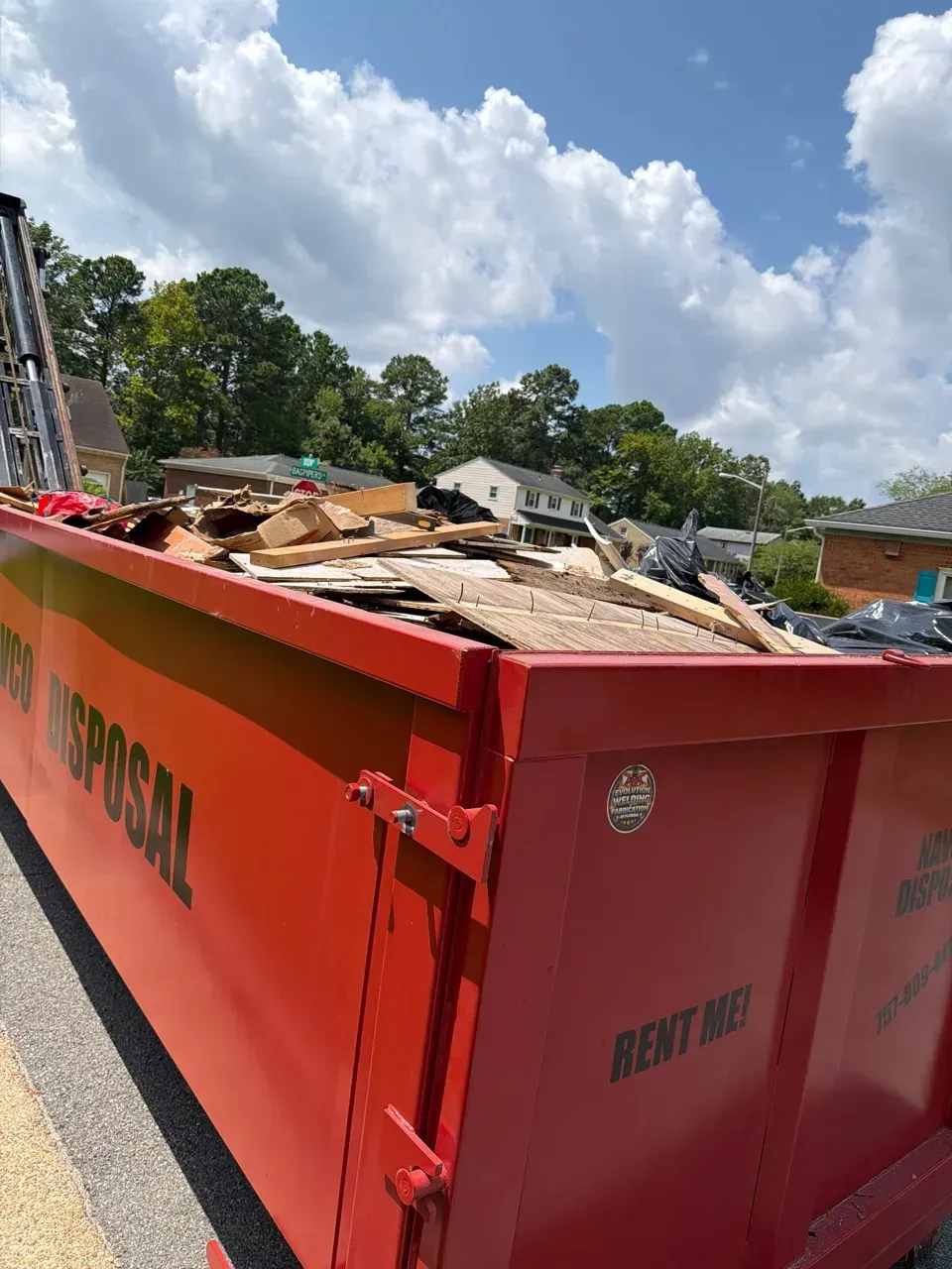 Large red metal dumpster filled with construction debris, sitting on grass near houses.