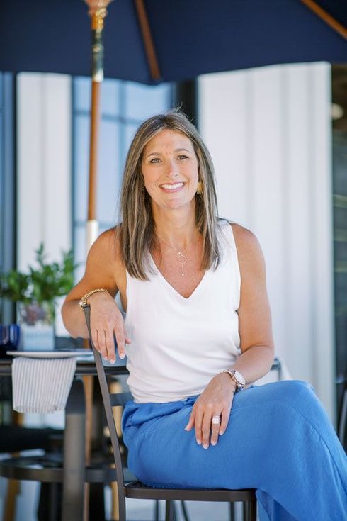 Woman seated under a blue canopy, smiling in a white top and blue pants.