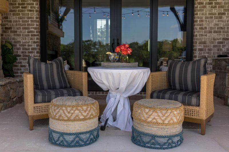 Patio seating area with two wicker chairs, round table with red flowers, and woven stools by glass doors