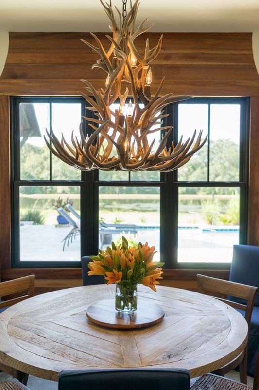 Rustic dining room with antler chandelier, round wooden table, and vase of orange flowers by large windows