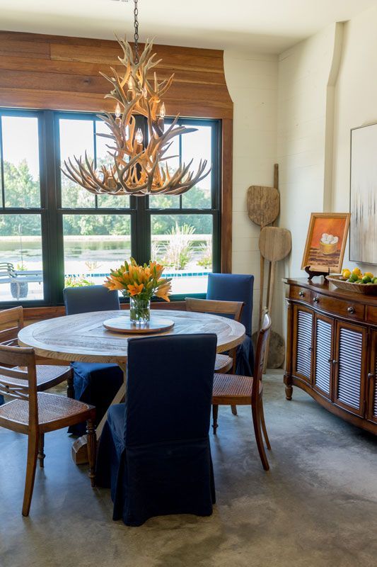 Dining room with round table, blue chairs, chandelier, and large window overlooking greenery.