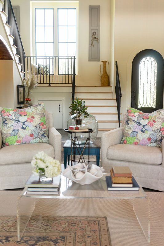 Bright living room with cream sofas, floral pillows, glass coffee table, and staircase by tall windows