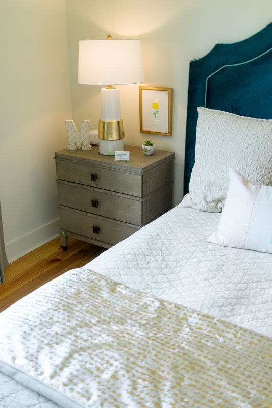 Cozy bedroom corner with a gray nightstand, lamp, framed art, and a bed with blue upholstered headboard.