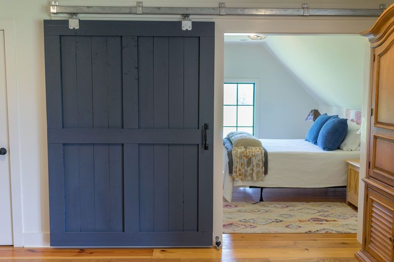 Dark gray barn door opens to a bright bedroom with a bed, blue pillows, and wooden floor.