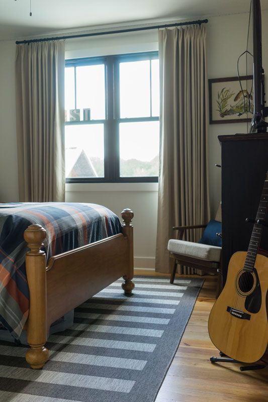 Bedroom with wooden bed, striped rug, large window, chair, and acoustic guitar leaning against the wall