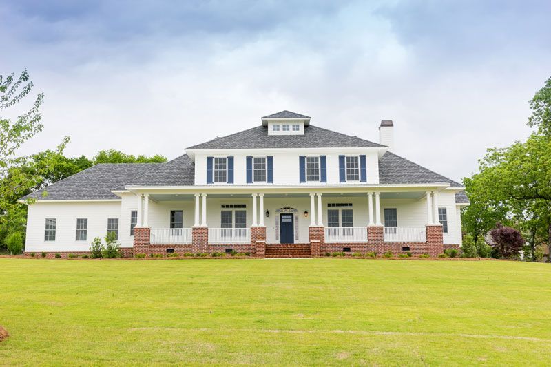 Large white farmhouse with a front porch, red-brick columns, and a wide green lawn under a cloudy sky