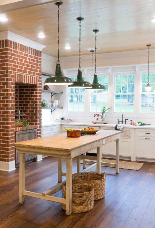 Bright farmhouse kitchen with a wooden island, hanging lamps, brick fireplace, and white cabinets near a window.