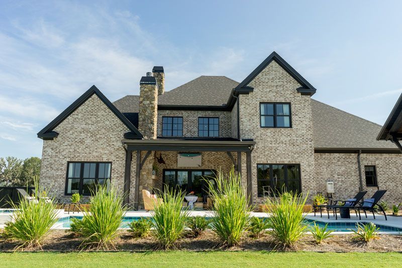 Large stone suburban house with black-trim windows, front lawn, and patio chairs under a bright sky