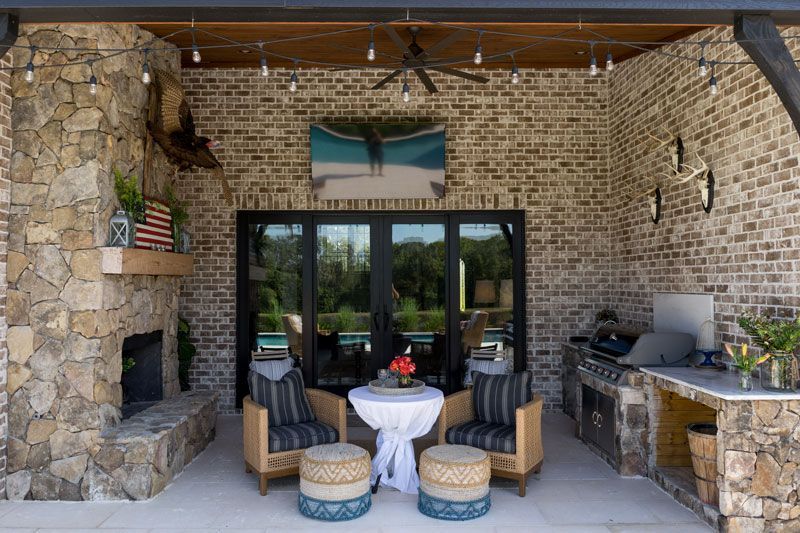 Stone patio with covered seating area, wicker chairs, table, built-in grill, and glass doors overlooking greenery