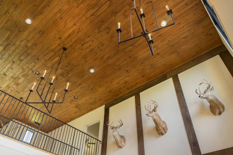 Wood-paneled vaulted ceiling with black chandeliers and mounted deer head artwork in a modern lodge interior