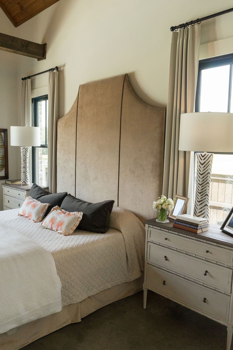 Elegant bedroom with a beige upholstered bed, white dresser, lamps, and tall windows with curtains