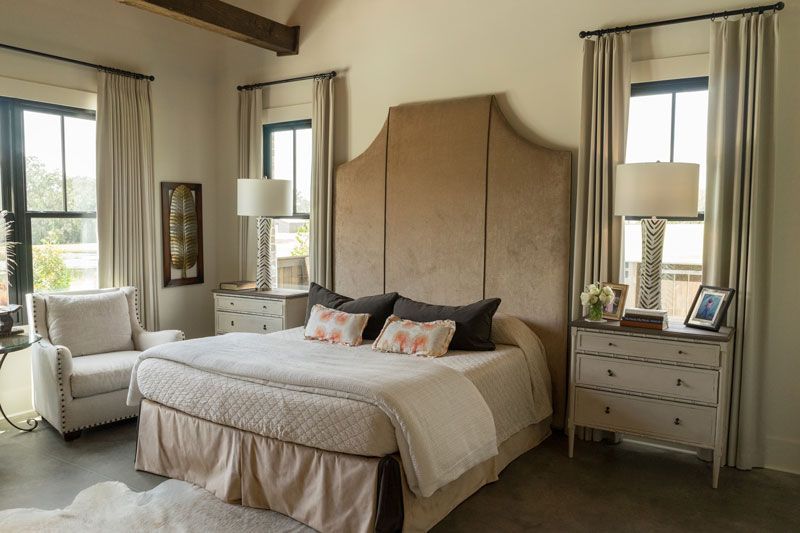 Bright bedroom with a large bed, tall beige headboard, and windows on both sides, with white nightstands and lamps.