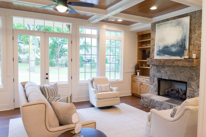 Bright living room with beige chairs, stone fireplace, and large windows overlooking a green yard