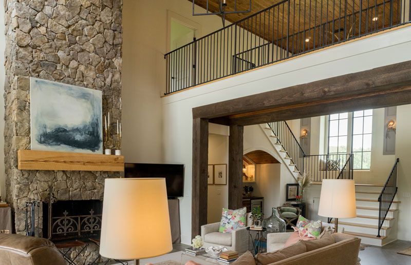 Rustic living room with stone fireplace, loft railing, staircase, and large windows glowing with warm light