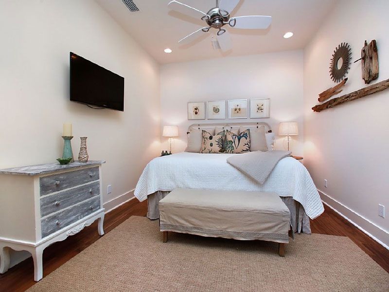 Small bedroom with white bed, wall-mounted TV, dresser, and ceiling fan in a bright neutral decor.