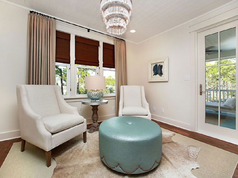 Bright living room with two white chairs, teal ottoman, chandelier, and window seating area