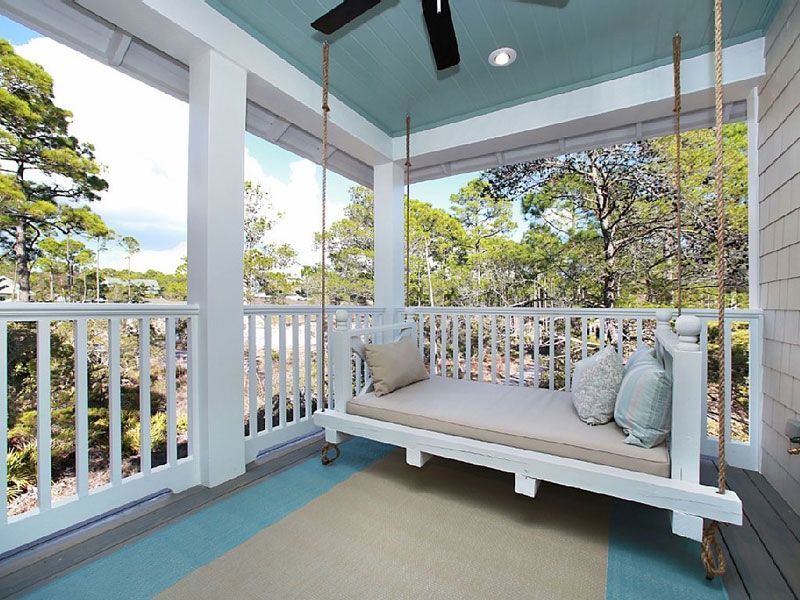 White porch swing on a covered patio with railings, teal ceiling, and leafy trees beyond