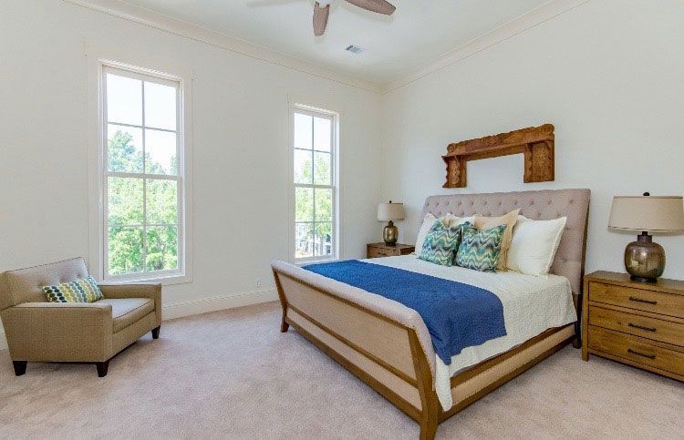 Bright bedroom with a wooden bed, blue blanket, two tall windows, and a beige chair by the wall