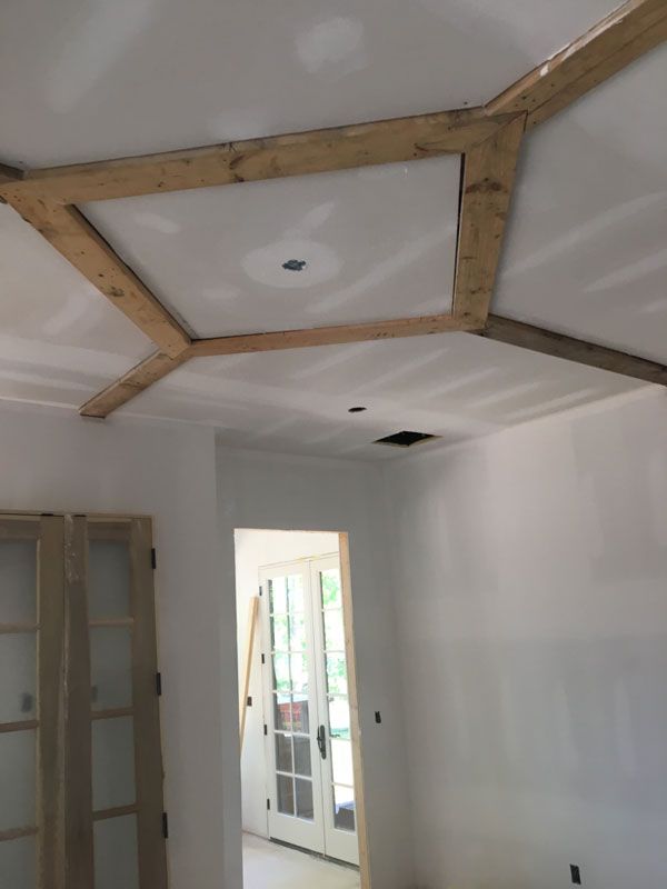 Renovated room with wood-framed ceiling panel and white walls, doorway to a brighter adjoining space.