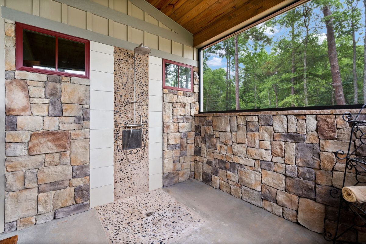 Stone patio under a covered porch with a white wall, red-trimmed window, and wooded view beyond.