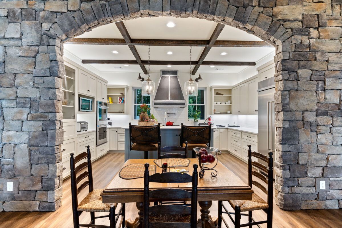 Stone arch framing a modern kitchen and dining area with white cabinets, wood table, and exposed beam ceiling