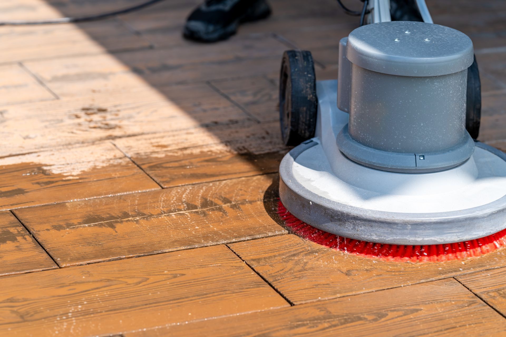 Floor buffer polishing a wooden floor, with a red scrubbing pad and a person’s shoe visible nearby