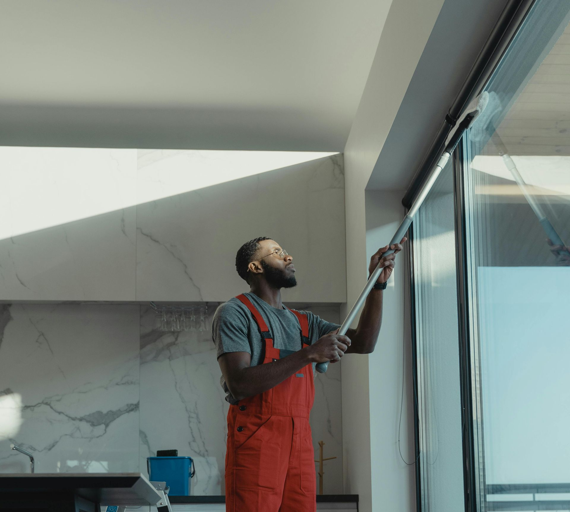 Person in red overalls cleaning large window with long-handled squeegee. Interior shot with sunlight.