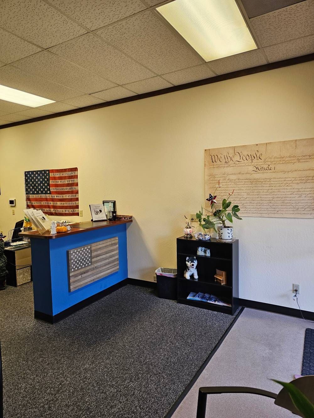 Office reception area with blue desk, American flag, black cabinet, and wall art under fluorescent lights