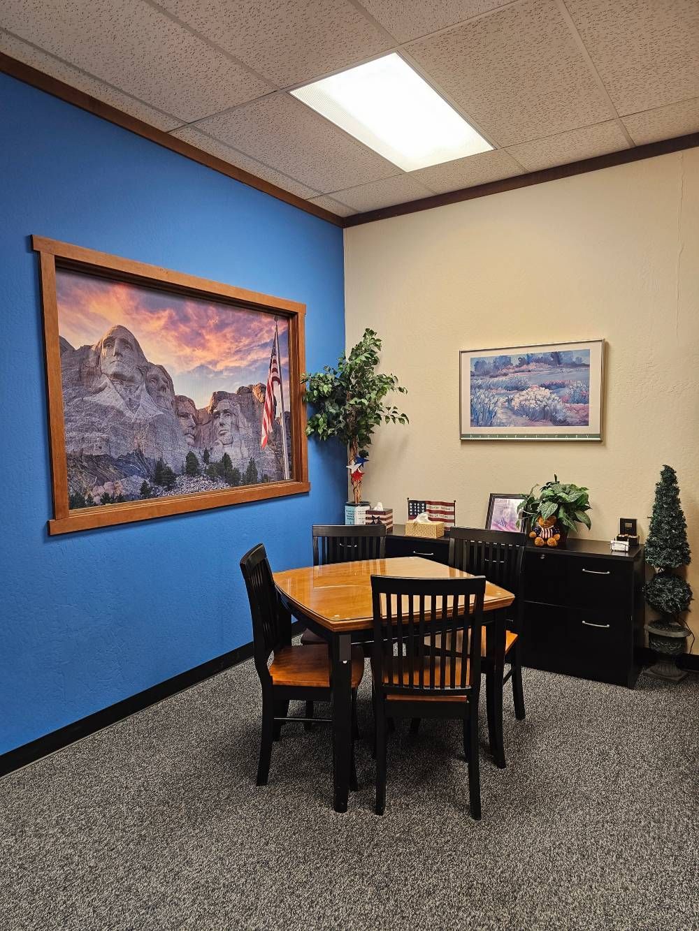 Small office meeting room with blue accent wall, table, chairs, framed art, plants, and desk.