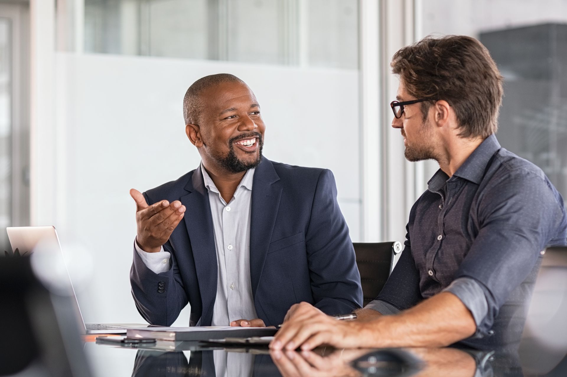 Two colleagues in a meeting, smiling and gesturing at a desk in a bright office