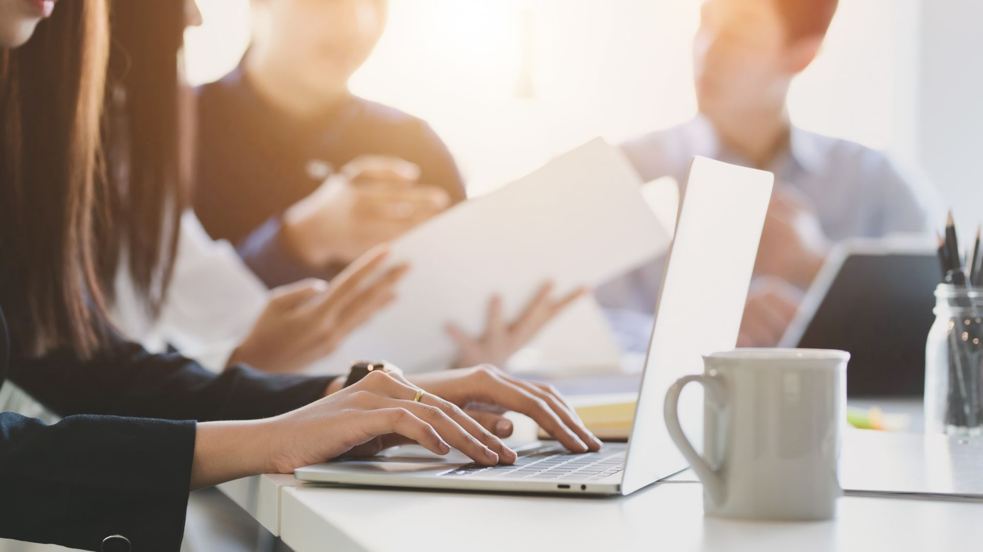 People working at a conference table with laptops and coffee mugs in a bright office.
