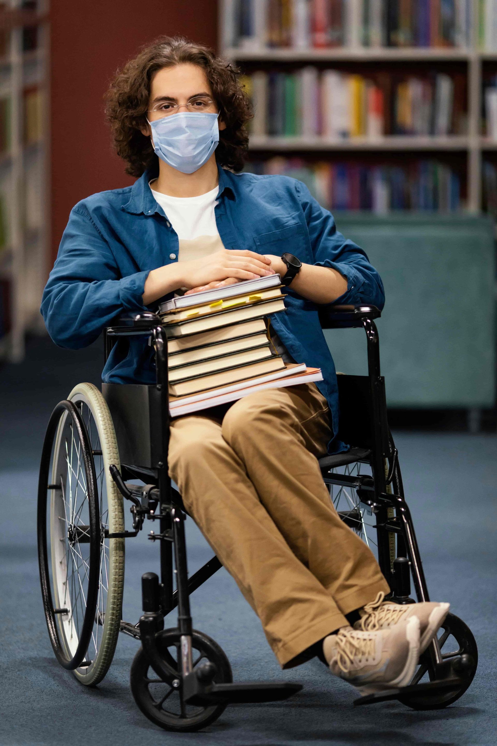 Person in a wheelchair holding books in a library, wearing a face mask.