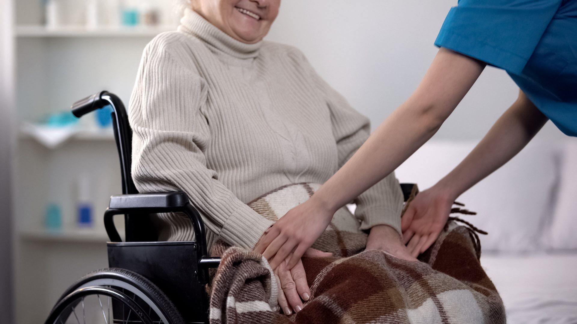 Smiling seated person in a wheelchair receiving a blanket from a caregiver in a home setting