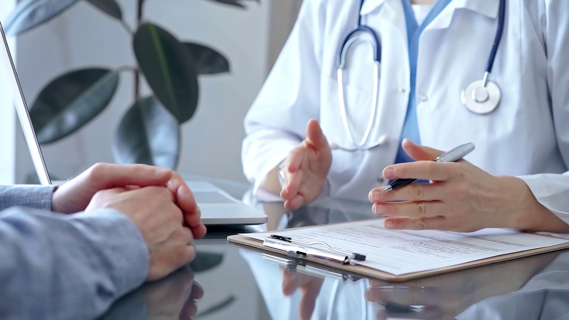A medical professional in a white coat talks to a patient across a desk, gesturing toward a clipboard.