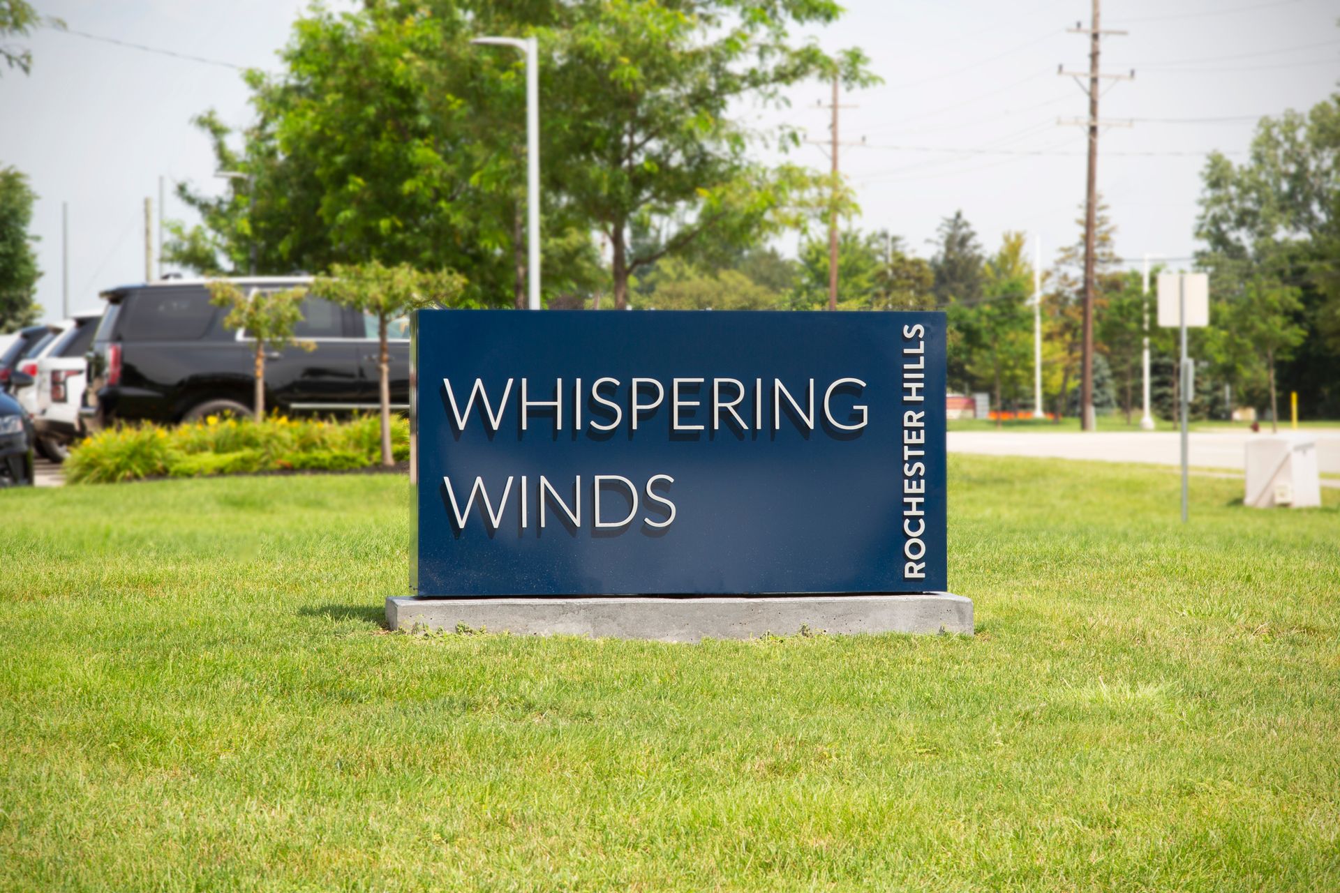 A blue sign that says whispering winds is in the middle of a lush green field.