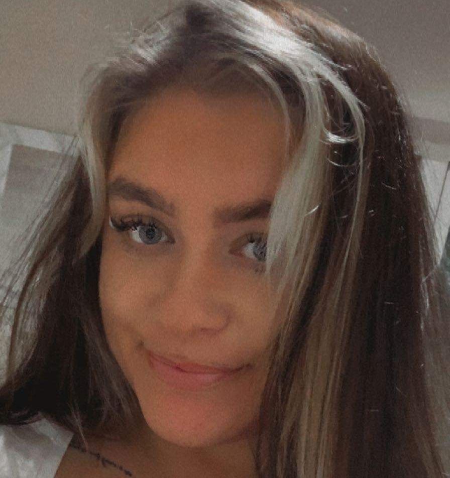 Close-up selfie of a person with long brown hair and a slight smile indoors