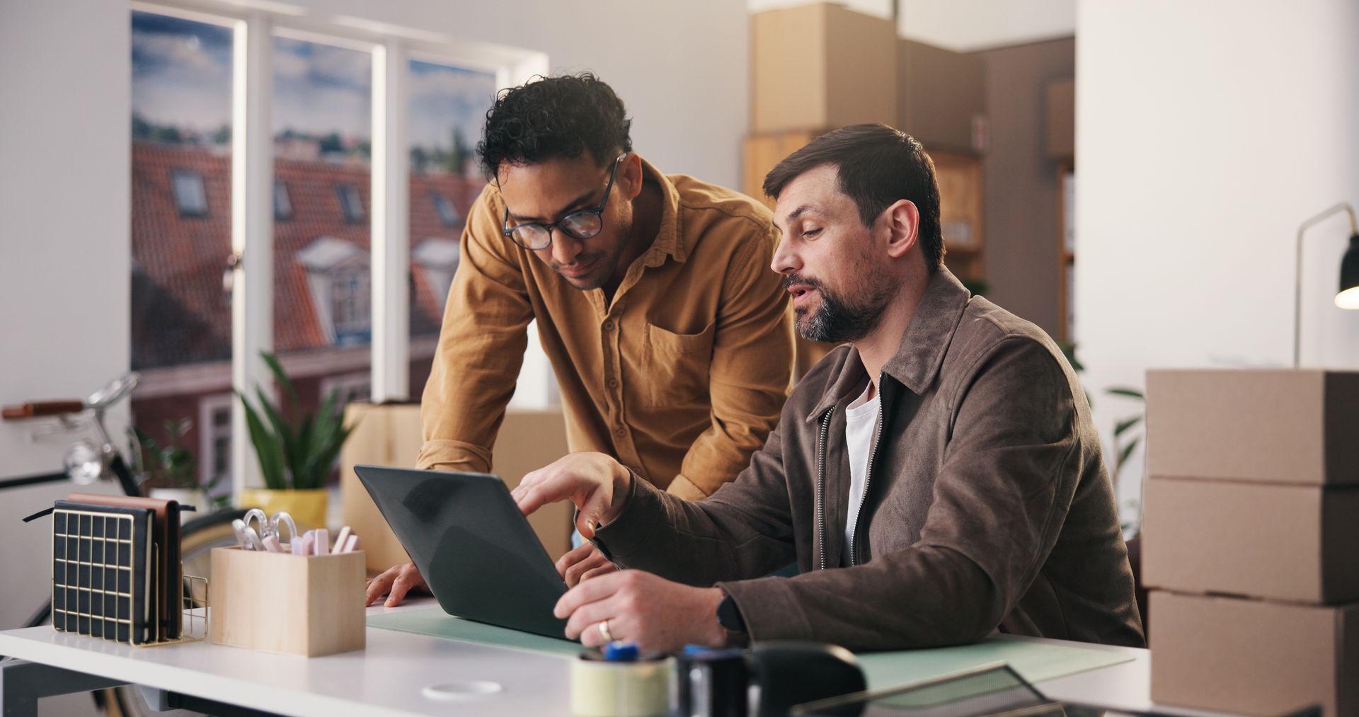 Two colleagues reviewing a laptop at a desk in a bright office, with stacked boxes nearby. Two colleagues reviewing a laptop at a desk in a bright office, with stacked boxes nearby.