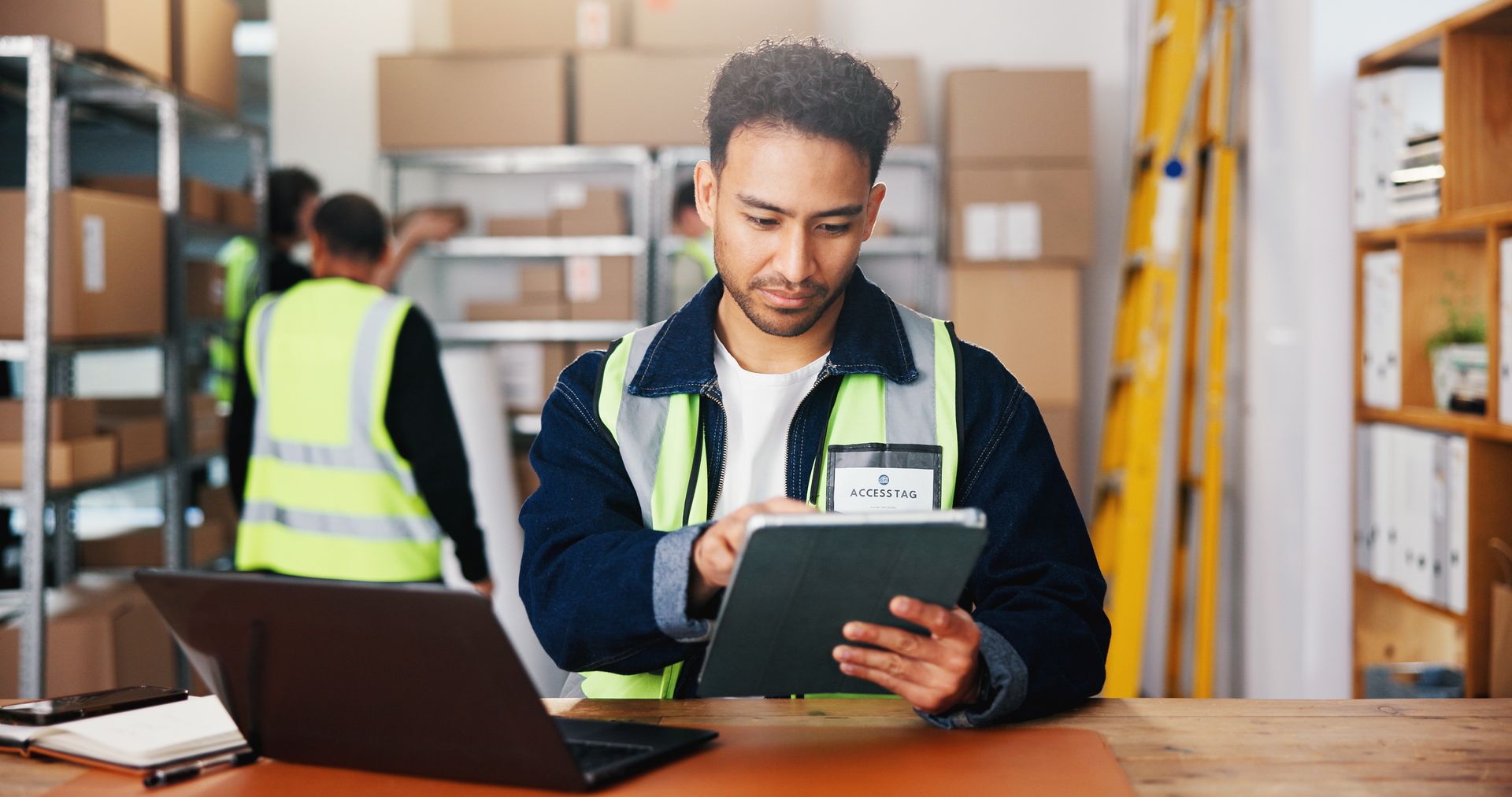 Warehouse worker in a safety vest using a tablet beside a laptop in a storage aisle Warehouse worker in a safety vest using a tablet beside a laptop in a storage aisle